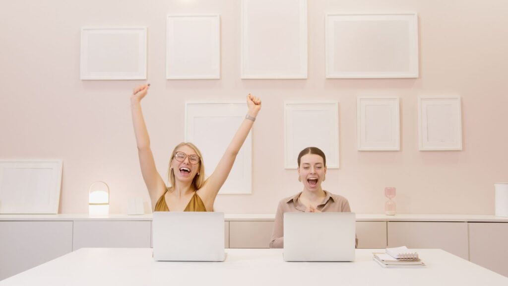 two girls working remotely using laptops, celebrating a work achievement.